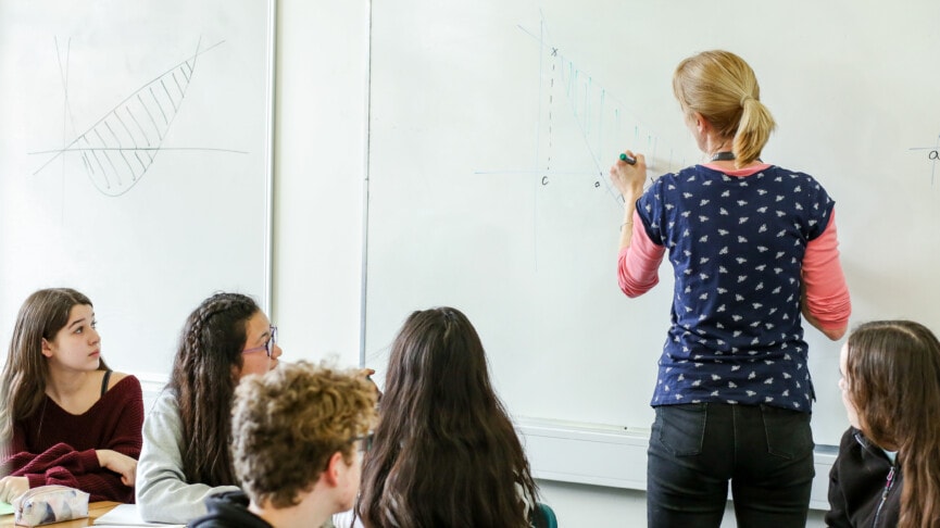 Maths teacher writing on white board with class watching